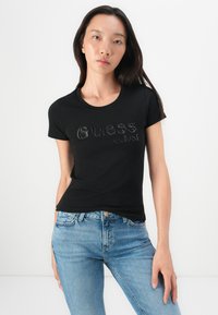 Black cotton t-shirt with a round neckline, featuring 'Guess' logo in shiny embellishments and year '1981' beneath it. Fitted design.