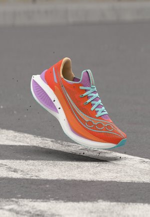 Zapatilla de running de color naranja brillante con detalles en púrpura y turquesa, en el aire sobre una superficie pavimentada con rayas blancas, con pequeños escombros visibles debajo.