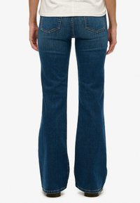 Jeans de mezclilla azul oscuro de campana con bolsillos traseros, cintura de tiro medio y costuras visibles. Textura suave, diseño clásico de cinco bolsillos.