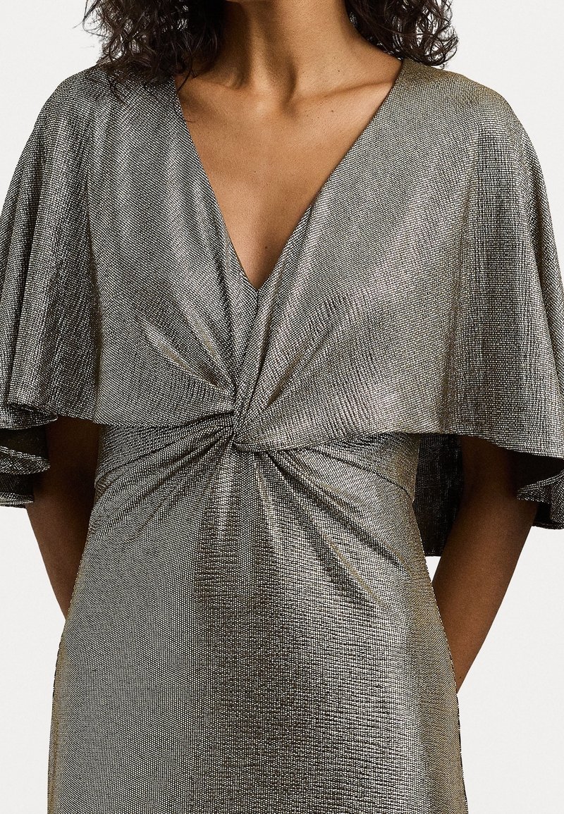 Metalická stříbrná šaty s hlubokým V výstřihem, lehkými rukávy a překrouceným uzlem v pase. Texturovaná tkanina odráží světlo.