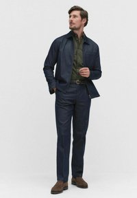 Veste en denim foncé avec un col pointu, chemise olive à boutons en dessous, associée à un pantalon foncé et des chaussures en daim marron.