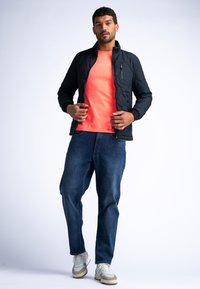 Schwarze Steppjacke mit hohem Kragen, leuchtendes oranges T-Shirt, blaue Jeans und grau-weiße Sneaker in stehender Pose.
