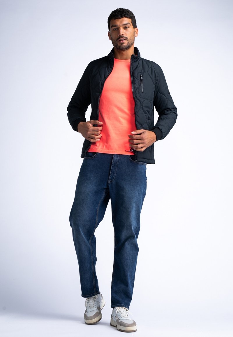 Schwarze Steppjacke mit hohem Kragen, leuchtendes oranges T-Shirt, blaue Jeans und grau-weiße Sneaker in stehender Pose.