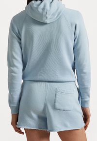 Henkilö, joka on pukeutunut vaaleansiniseen huppariin ja vastaaviin risarajaisiin shortseihin, näkyy selkäpuolelta yksinkertaista taustaa vasten.