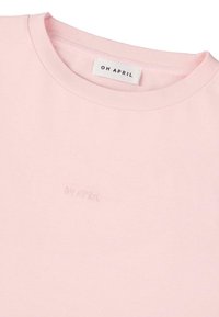 T-shirt en coton rose clair avec un col rond, arborant un logo brodé "OH APRIL" dans une couleur assortie. Texture lisse.