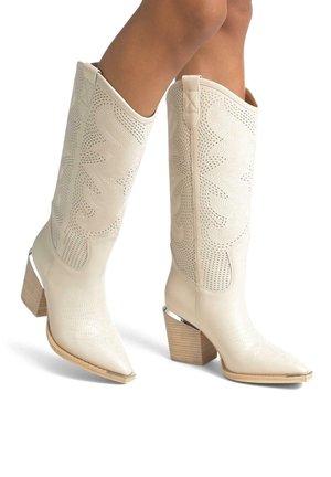 Bottes de cowboy en cuir beige hauteur mollet avec motifs cousus, talons blocs en bois et bouts métalliques portées sur des jambes nues.