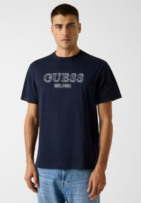 Marineblaue Baumwoll-T-Shirt mit "GUESS"-Logo und "EST. 1981" in kontrastierender Stickerei. Kurze Ärmel, Rundhalsausschnitt, lockere Passform.