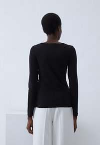 Svart langermet topp med rund hals, laget av et glatt, strekkbart stoff. Har en slim fit og rekker til livet, kombinert med hvite bukser.