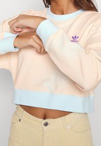 Felpa corta in morbido tessuto pesca con polsini e orlo blu chiaro. Presenta un piccolo logo Adidas viola sul petto. Elementi di design testurizzati.