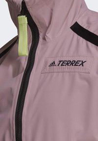 Terrex regnjacka, lätt rosa tyg, med en svart dragkedja och detaljer, samt en grön dragflik och en logotypapplikation på bröstet.