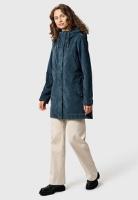 Nainen kiharat hiukset, päällään tummansininen hupullinen takki, beiget housut ja mustat saappaat, seisoo yksinkertaista vaaleanharmaata taustaa vasten.