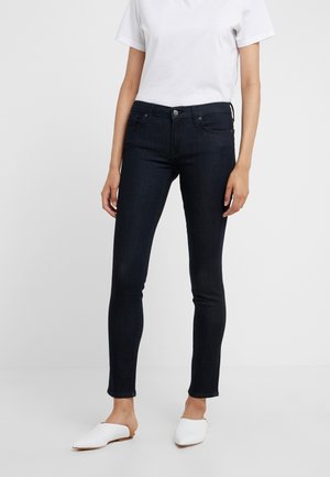 Mujer con vaqueros ajustados oscuros, camiseta blanca de manga corta y zapatos blancos sin cordones, de pie contra un fondo claro y liso.