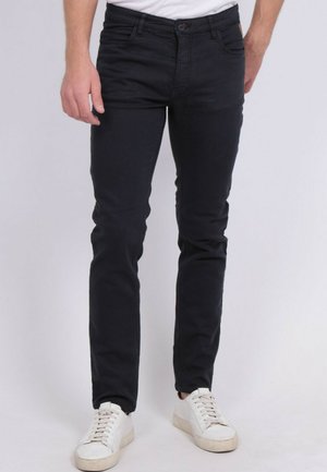 Homme portant un jean noir slim et des baskets blanches, debout devant un fond uni.