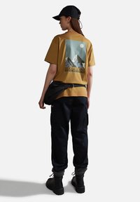 Camiseta marrón con gráfico de montaña, pantalones cargo negros, calcetines texturizados y botas negras. Incluye una bolsa de hombro y una gorra.
