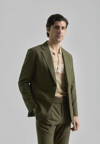 Traje entallado en verde olivo con un pantalón a juego. Debajo, una blusa beige con un patrón texturizado y escote abotonado.