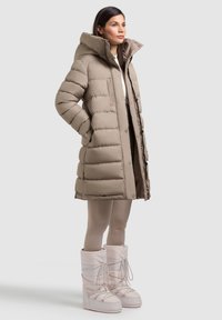 Béžová vatovaná bunda s vysokým golierom, zapínaním na cvoky a bočnými vreckami. Nosená s béžovými pančuchami a bielymi zimnými topánkami so šn�úrkami.