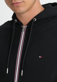 Un sweat à capuche noir avec fermeture éclair argentée et accents à rayures rouges. Il comporte un petit logo sur la poitrine et des cordons pour la capuche.