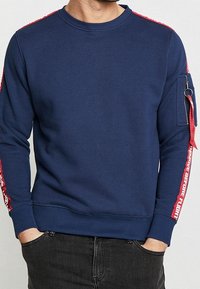 Marinblå sweatshirt med röda detaljer på ärmarna, med en ficka med dragkedja och textdetaljer längs sidan. Mjuk tyg.