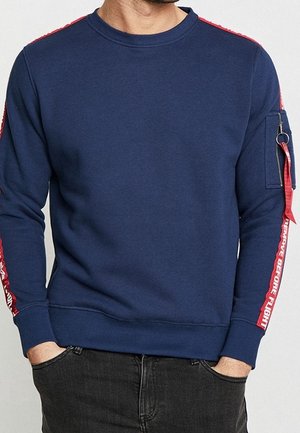 Sweat-shirt bleu marine avec des accents rouges sur les manches, doté d'une poche patch avec fermeture éclair et d'un texte le long du côté. Tissu doux.