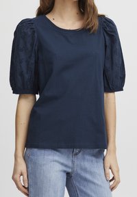T-shirt en coton marine avec manches courtes bouffantes ornées de motifs floraux brodés. Encolure ronde et coupe décontractée, associée à un jean clair.