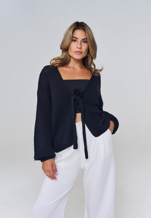 Mujer con cabello rubio y ondulado que lleva un cárdigan de punto negro con nudo frontal sobre una camiseta negra y pantalones blancos de pierna ancha, de pie frente a un fondo liso.