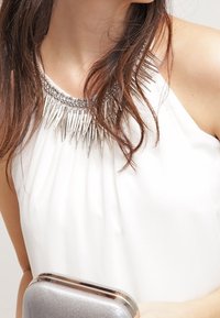 Vit ärmlös topp med veckat tyg och ett silverfärgat, taggigt halsband. Modellen håller en silverglittrig kuvertväska.