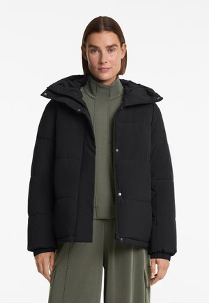 Schwarze Pufferjacke mit Kapuze, ausgestattet mit einem gesteppten Design und Druckknöpfen. Über einem grünen Oberteil mit hohem Kragen getragen. Glatte Textur, lockere Passform.