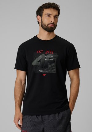 Mężczyzna z krótkimi kręconymi włosami i brodą, ubrany w czarną koszulkę z czerwonym i białym napisem "EST 1922" oraz graficznym wzorem z przodu.