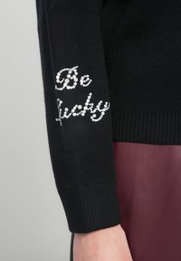 Melns džemperis ar ribotām aprocēm, kas piedāvā uzrakstu "Be Lucky" uz piedurknes, izrotātu ar rhinestones. Gluda tekstūra, cieši pieguļošs dizains, kontrastēts ar bordo audumu.