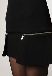 Jupe noire avec un ourlet évasé, dotée de deux accents de fermeture éclair en argent sur les côtés. Fabriquée en tissu doux avec une texture lisse.