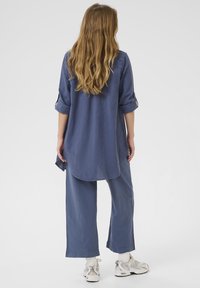Blue, lightweight linen shirt with rolled-up sleeves, curved hemline, paired with matching blue wide-leg pants and white athletic shoes.