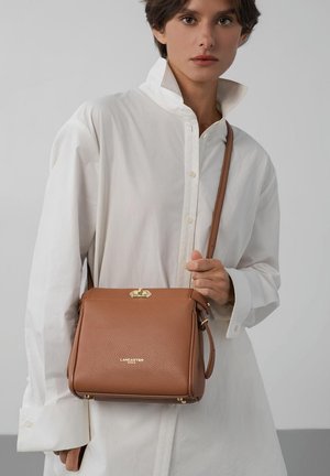 Woman wearing oversized white shirt holds brown leather Lancaster shoulder bag with gold clasp against gray background.