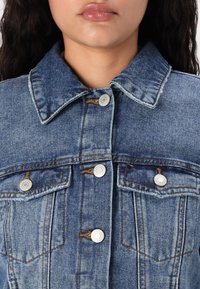 Chaqueta de mezclilla en azul oscuro, con cuello clásico, botones plateados y dos bolsillos en el pecho con detalles de costura. Tejido texturizado con áreas deslavadas.