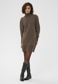 Robe en tricot marron à col roulé avec une coupe décontractée, des poignets et un ourlet côtelés. Portée avec des bottes en cuir noir arrivant au genou.