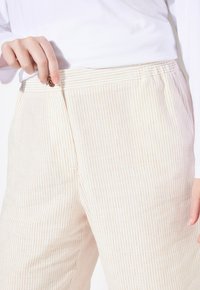 Béžové a krémové pruhované kalhoty s elastickým pasem a předním zipem, se zdobným kovovým knoflíkem. Hladká textura látky.