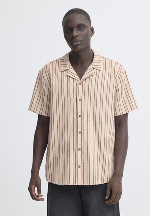 Joven con camisa beige de manga corta con rayas rojas verticales, de pie contra un fondo blanco liso.