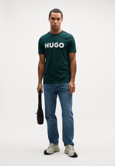Зелена памучна тениска с надпис "HUGO" в кремаво, съчетана с сини дънки и бежови спортни обувки. Черен чанта през тялото в ръка.