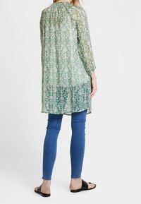 Grön gradient tunika med intrikat vitt blommönster, långa ärmar, rund halsringning, lätt tyg, parat med blå jeans och sandaler.