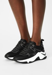 Baskets noires en matériau mesh et cuir, avec des lacets réfléchissants, une semelle blanche et des accents de logo sur la languette et le côté.