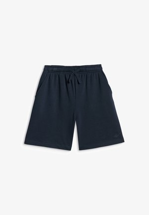 Garçon debout, les mains dans les poches, portant un t-shirt vert, un sweat à capuche zippé bleu marine, un short bleu marine, des chaussettes blanches et des baskets blanches sur un fond uni.