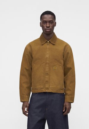 Uomo che indossa una giacca marrone con cerniera e due tasche frontali e pantaloni blu scuro, in piedi davanti a uno sfondo chiaro e uniforme.