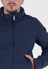 Marineblaue Zip-Jacke aus weichem Material mit Kapuze, ausgestattet mit einem durchgehenden Reißverschluss und Seitentaschen, ergänzt durch ein Logo auf dem Ärmel.