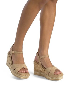 Beige suede wedge sandals with a woven jute sole, featuring crisscross straps and an adjustable ankle strap. Open-toe design.