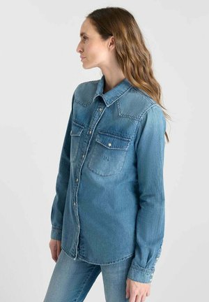 Vrouw die naar links kijkt, gekleed in een vervaagd blauw denim overhemd met knoopjes op de borstzakken en bijpassende blauwe jeans voor een effen achtergrond.