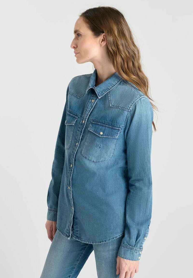 Kvinde, der står til venstre iført en falmet blå denimskjorte med knaplukket brystlommer og matchende blå jeans foran en ensfarvet baggrund.