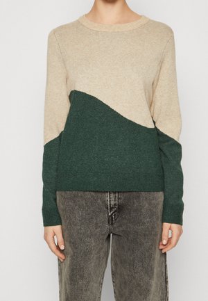 Personne portant un pull beige et vert foncé en bloc de couleur avec des manches longues et un jean gris foncé délavé devant un fond uni.