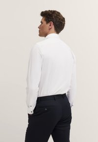 Weißes Langarmhemd mit klassischem Kragen, kombiniert mit schwarzen Hosen mit geknüpften Gesäßtaschen. Glattes Material, taillierte Passform.