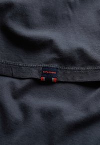 Tejido azul marino oscuro con una textura suave; cuenta con una etiqueta de marca en rojo, azul y blanco etiquetada como "SUPERDRY" en la costura.