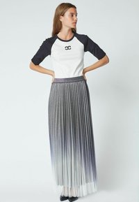 Un top raglan alb-negru cu un logo, asociat cu o fustă metalizată plisată de culoare gri care se estompează în alb. Modelul poartă încălțăminte neagră.