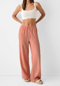 Femme portant un crop top en tricot blanc, un pantalon corail rose large avec cordon de serrage, et des sandales à plateforme beiges, se tenant devant un mur blanc uni.